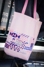 Pink tote bag with geometric design and text held by a person in an urban setting.
