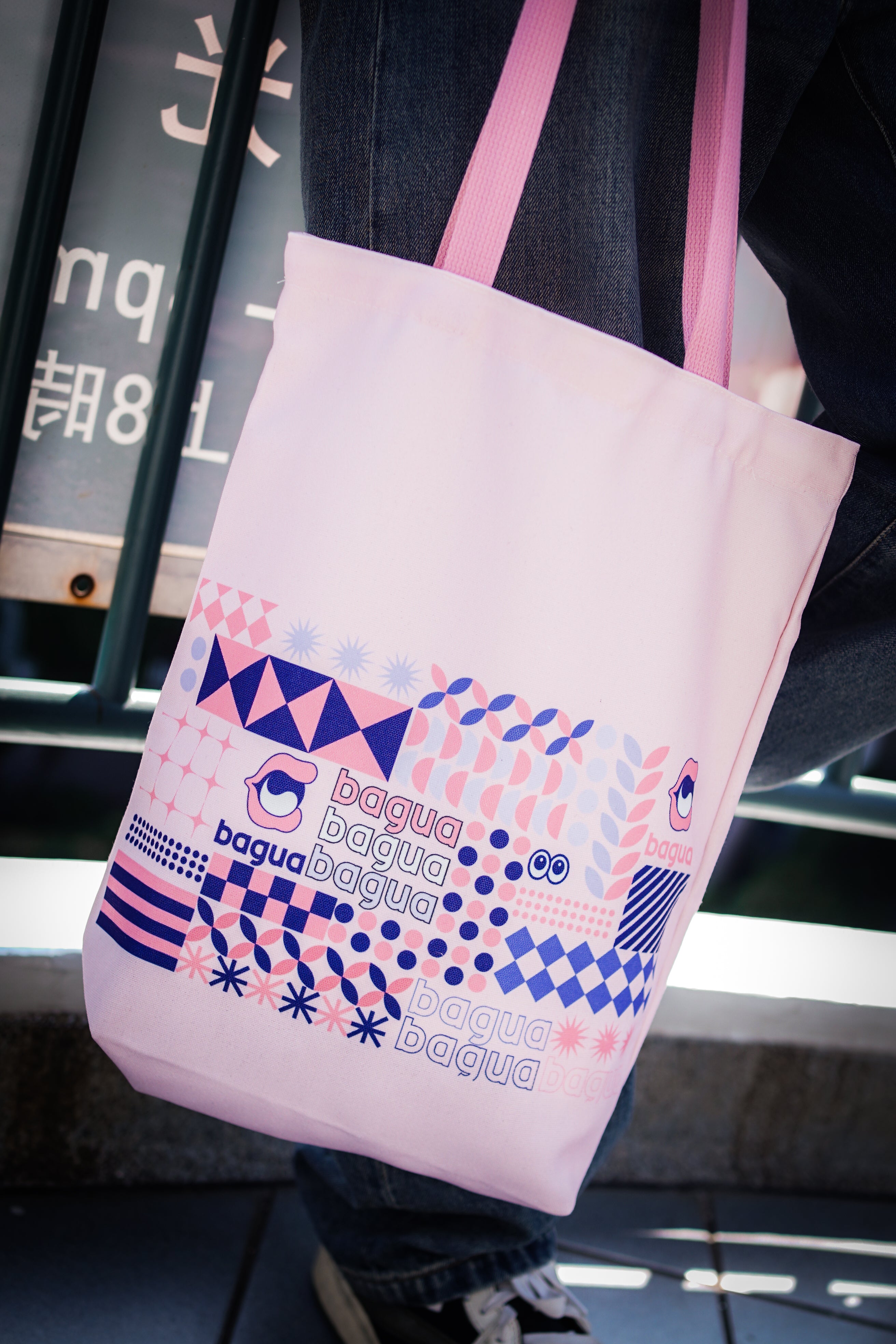Pink tote bag with geometric design and text held by a person in an urban setting.